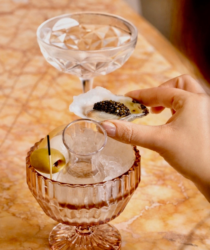 A hand holding a small bite topped with caviar beside two elegant cocktail glasses on a marble surface.