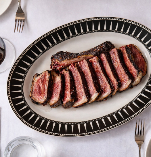 A sliced seared steak on a decorative black and white oval platter, served with a glass of red wine on a white tablecloth.