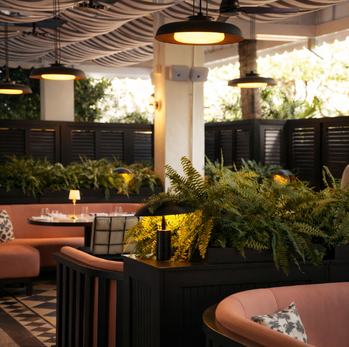 Modern outdoor restaurant patio with pink curved seating, black pendant lights, ferns, and striped canopy overhead