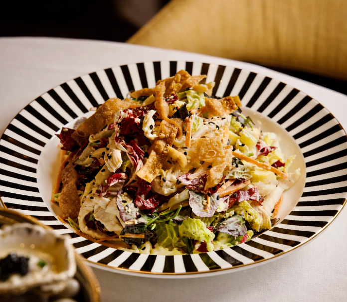 A chopped salad with radicchio, cabbage, crispy wontons, and sesame seeds in a black and white striped bowl on a white tablecloth.