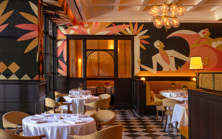 An elegant restaurant dining room with white-clothed tables, warm lighting, and bold Art Deco-inspired murals in orange, pink, and gold tones.