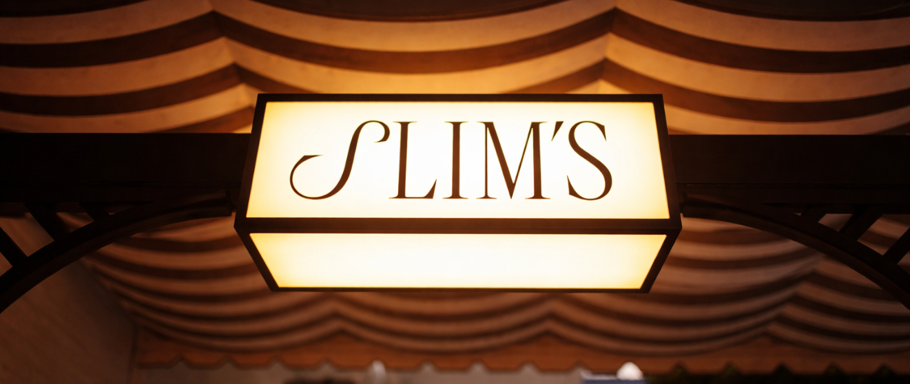 Illuminated rectangular sign reading "SLIM'S" in brown letters against decorative striped ceiling backdrop